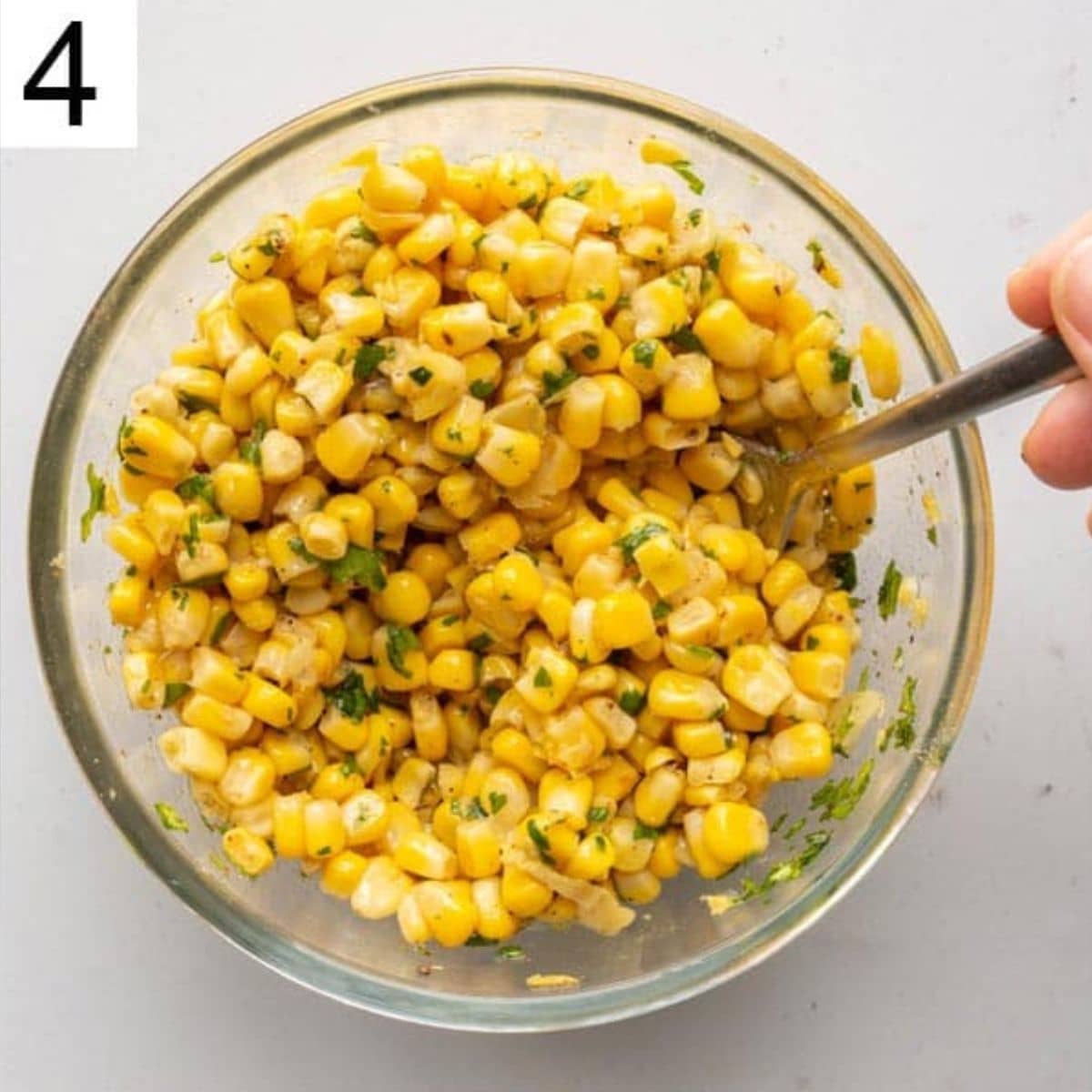 Sweetcorn and herbs being mixed in a glass bowl with a spoon and the number four in the upper left corner.