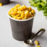 Small black cup wuth buttered yellow corn and herbs in it next to a spoon.