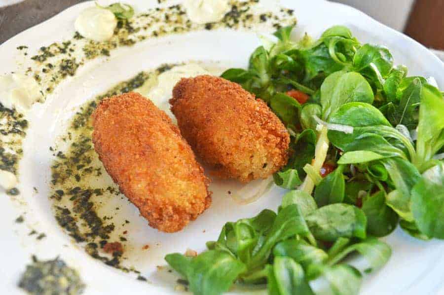 Two golden rice croquettes on a white plate with salad and aioli and pesto