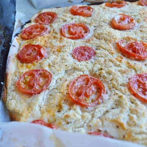 A side on view of a pizza with a white sauce and sliced tomatoes on a sheet of baking paper