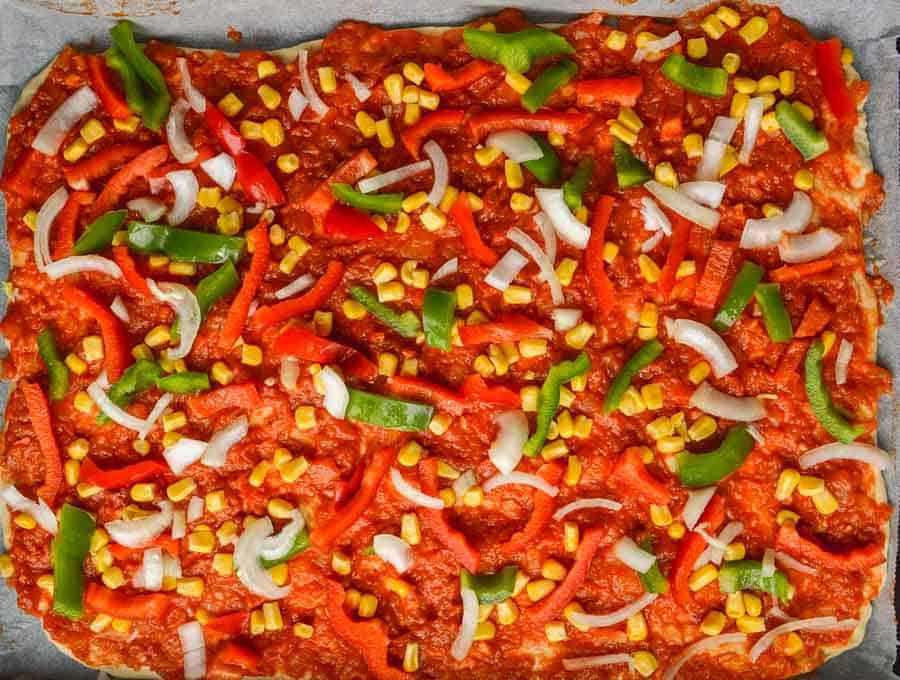 An uncooked pizza with tomato sauce, corn, peppers and onion on it