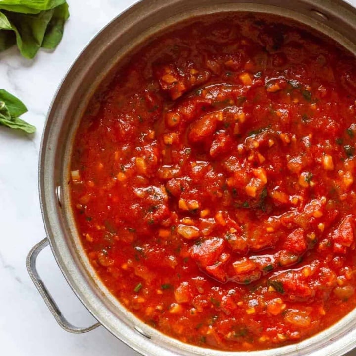 A silver pot of red tomato pasta sauce surrounded by fresh basil, tomatoes and pasta