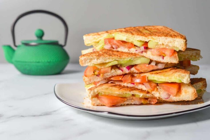A pile of tomato and cucumber masala sandwiches on a white plate with a green teapot in the background