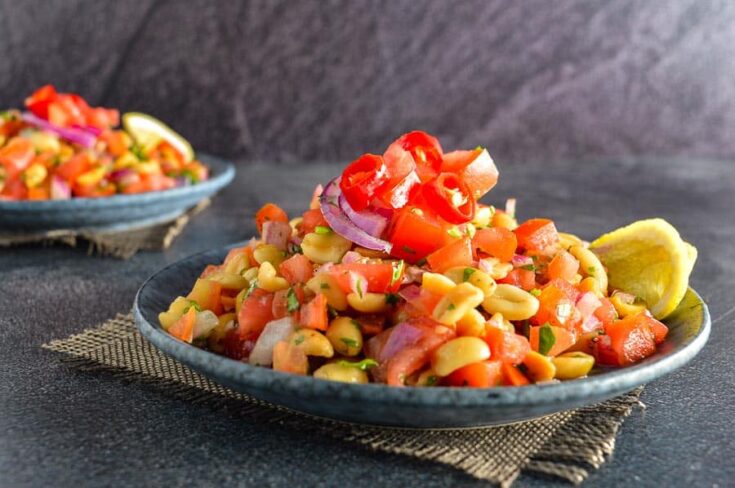 Closeup of a grey dish of peanuts, chili peppers, tomatoes, red onion and lemon slices