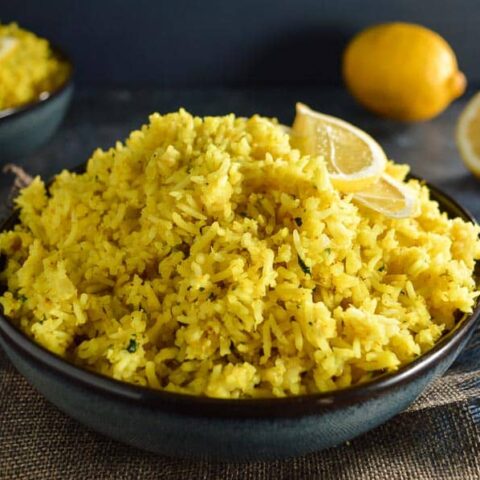 A dark blue bowl of yellow rich with 2 lemon slice halves and 2 lemons in the background