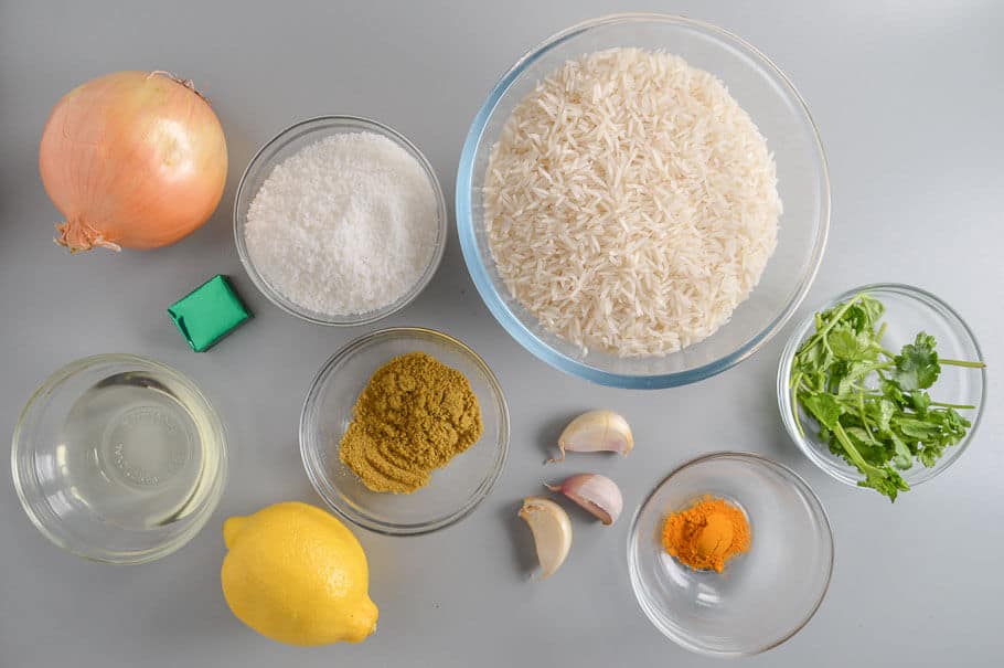 A lemon, an onion, 3 cloves of garlic, clear bowls of white rice, dessicated coconut, fresh cilantro, ground cilantro, turmeric and oil.
