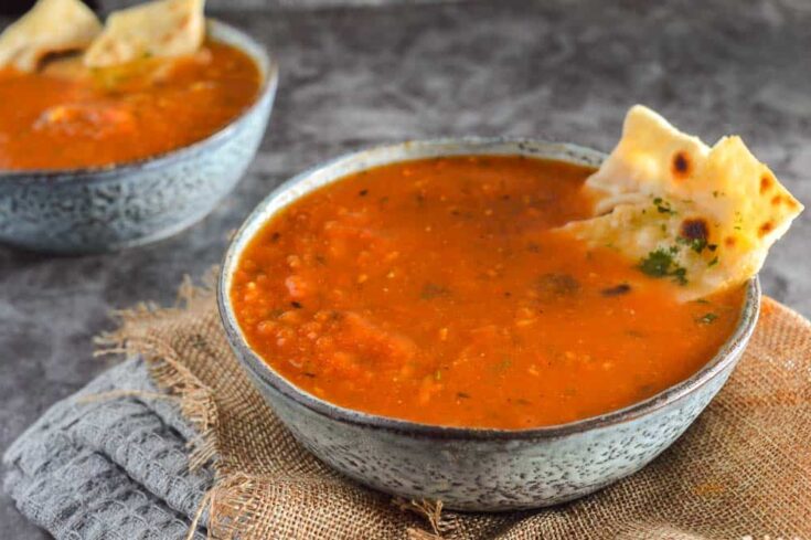 Two bowls of thick red soup with spices floating on top on a grey background