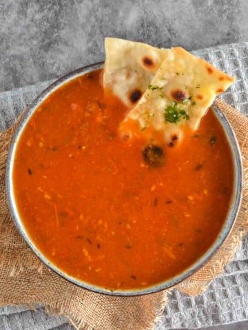 An overhead view of a bowl of red soup with two pieces of naan, on a grey background