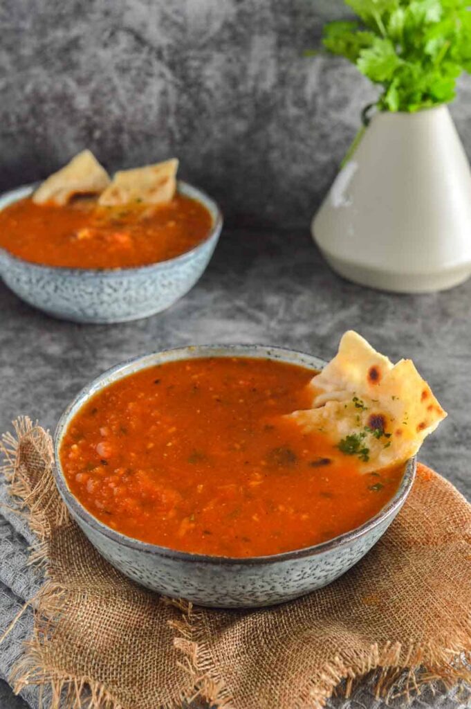 Two bowls of red soup with bread in it and a beige vase of fresh cilantro in the background