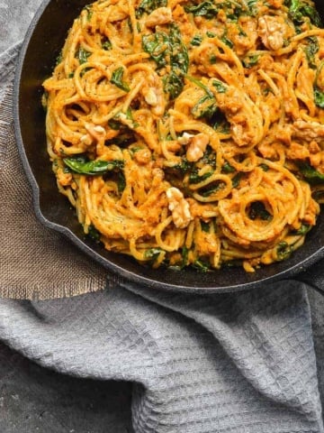 Top down view of pan full of orange spaghetti, walnuts and spinach on a grey teatowel
