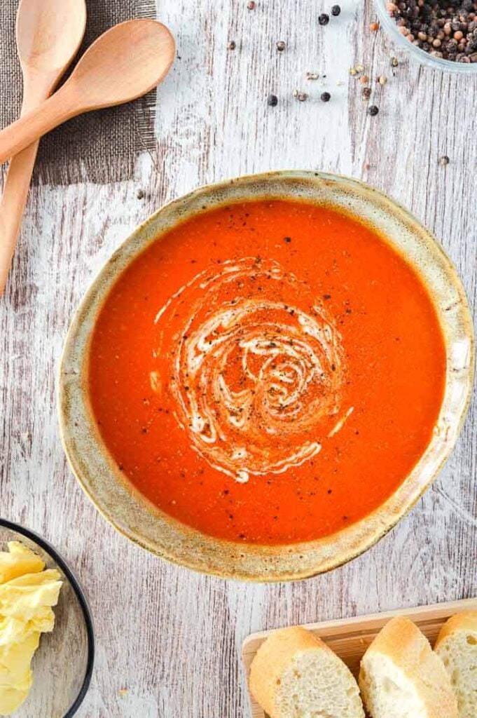 A bowl of tomato soup surrounded by wooden spoons and sliced baguette