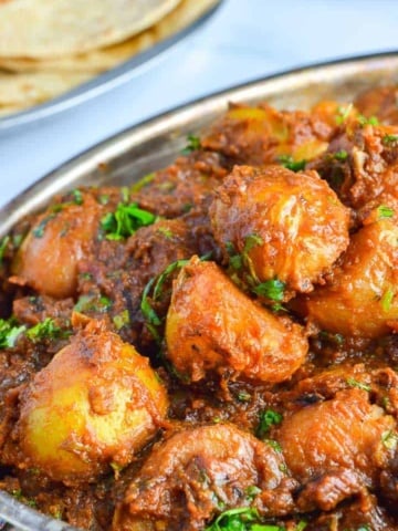 angled view of potatoes in brown sauce with fresh herbs sprinkled on top and roti in the background
