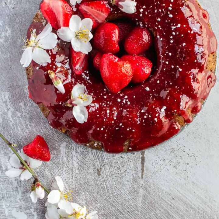 Top down image of a bundt cake glazed with a red fruit glaze and decorated with strawberries and white flowers on a grey background