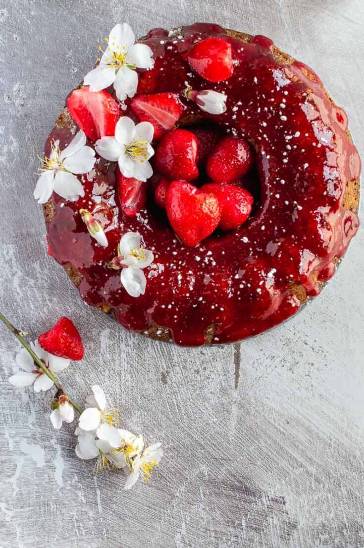 Top down image of a bundt cake glazed with a red fruit glaze and decorated with strawberries and white flowers on a grey background