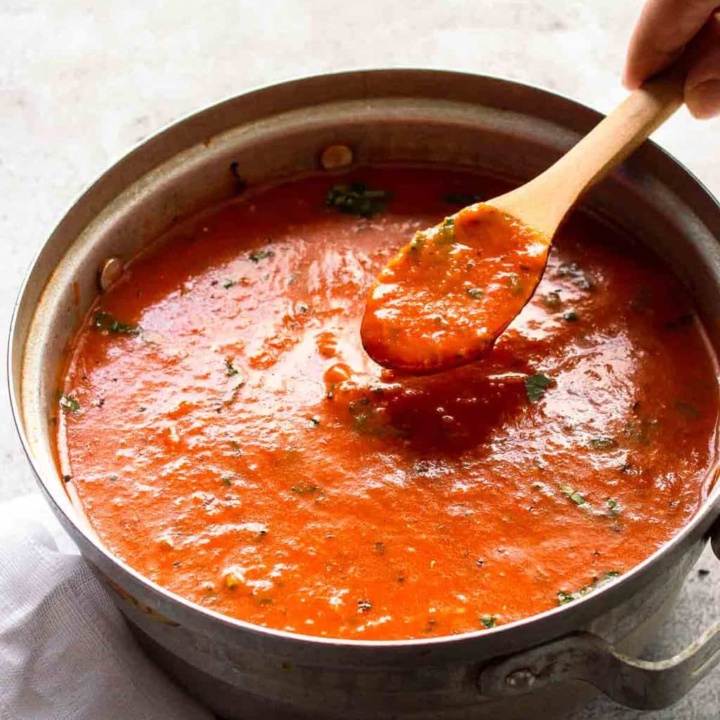 Thick reddy-orange sauce with herbs in a silver pot with a wooden spoon in it