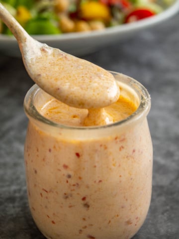 A silver spoon coated in creamy orange dressing over a small glass jar of it on a grey background.