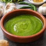 A brown bowl with green sauce on a dark grey backdrop with garlic and green chili peppers behind it.