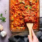 A spatula scooping up thick red sauce with chickpeas and vegetables from a glass casserole dish.