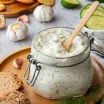 Creamy herbed white sauce in a jar surrounded by garlic, cucumbers, bread and crackers.