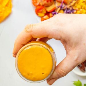 A hand holding a jar of orange sauce with yellow tortilla chips in the background.