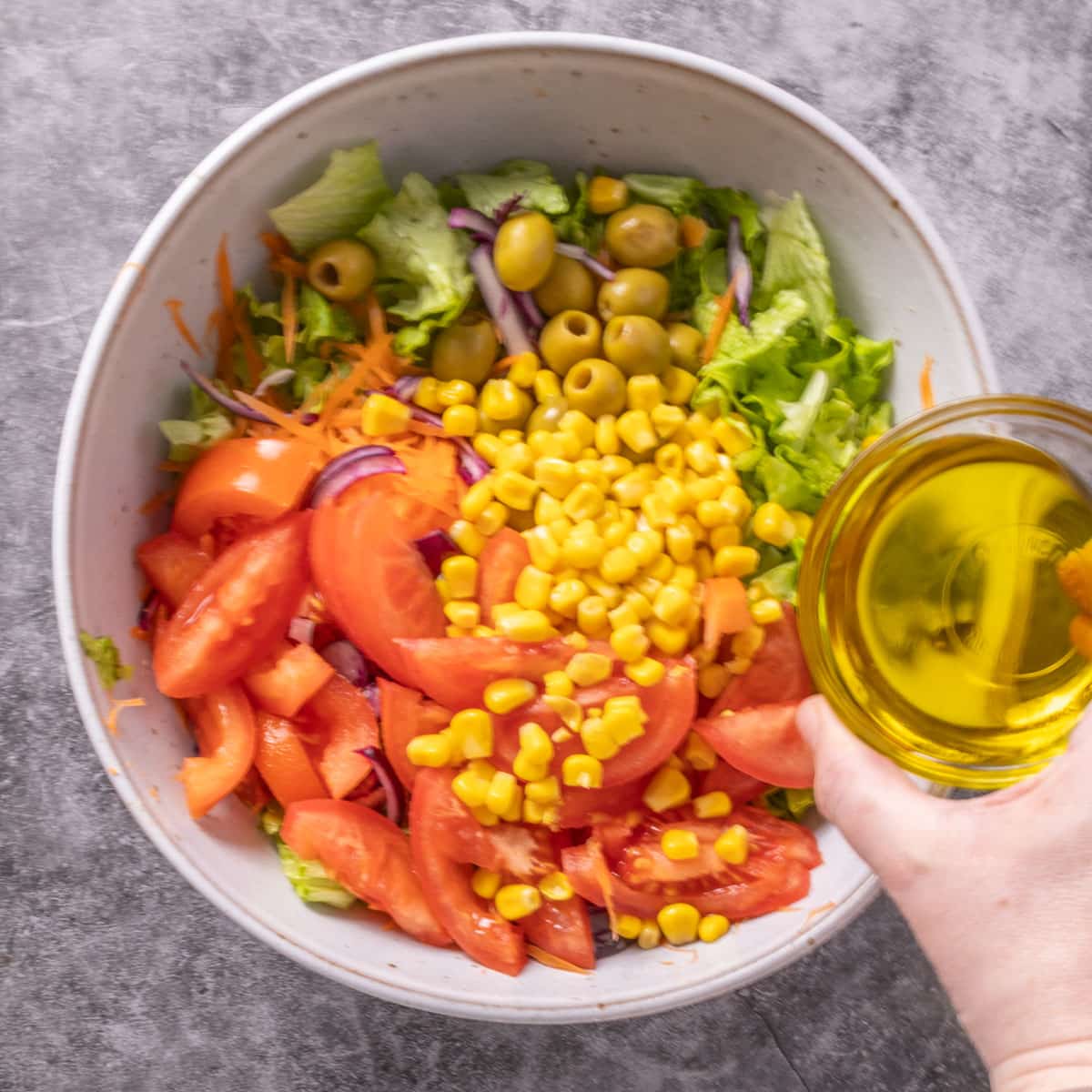 A grey bowl with corn, tomatoes, green olives and lettuce in it and yellow oil being poured in.