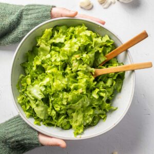A bowl of green salad with wooden spoons in it being held by a person wearing a green jumper.