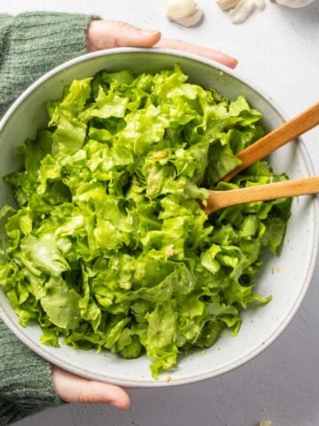 A bowl of green salad with wooden spoons in it being held by a person wearing a green jumper.