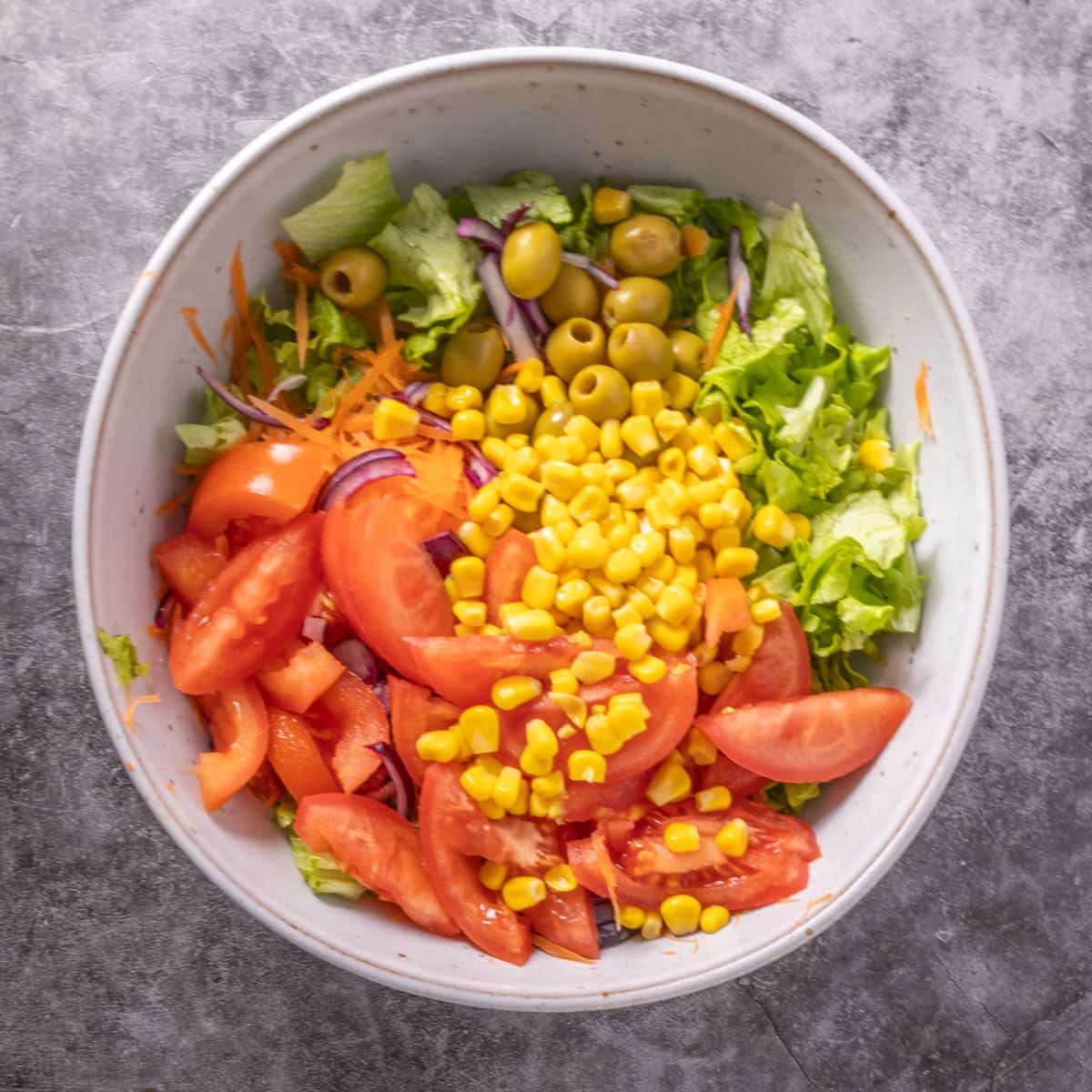 A grey bowl on a grey background with corn, sliced tomatoes, green olives and lettuce in it.