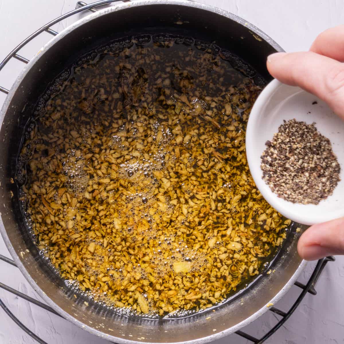 A pan with toasted garlic in oil and a hand adding a small white bowl of black pepper to it.