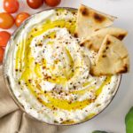 A bowl of thick white liquid with oil drizzled over accompanied by cherry tomatoes and pita chips.