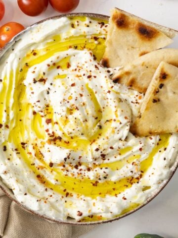 A bowl of thick white liquid with oil drizzled over accompanied by cherry tomatoes and pita chips.