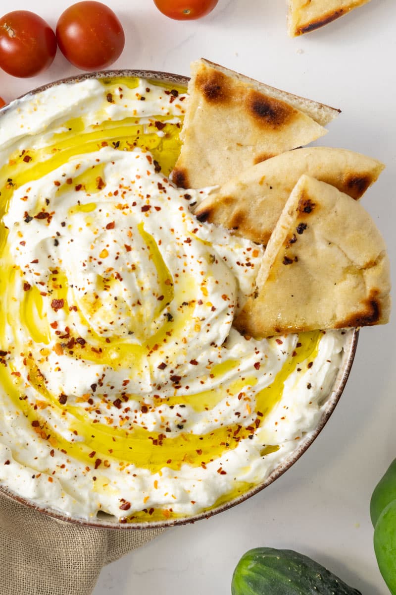 A bowl of thick white substance with oil drizzled on top and three pita chips dipped in it.